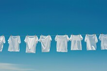 White T-shirts Hanging On The Line With Pegs On Clear Blue Sky Background. Many Simple White Shirts
