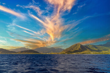 a beautiful spring landscape of the coast of Oahu with majestic mountain ranges, lush green trees and plants, rippling blue ocean water and blue sky with clouds at sunset in Kapolei Hawaii USA