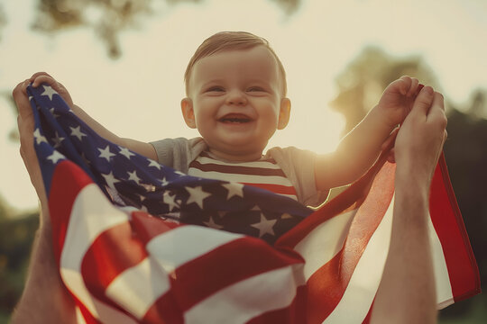 Smiling cute infant baby patriot on unrecognized parent`s hands holding America USA big flag draped over shoulders at sunny summer day. 4 July Independence Day celebration