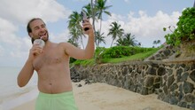 Unshaved Long-haired Macho Man With Bare Torso Capturing Selfie Sipping Drink From Glass, Surrounded By Restless Ocean, Exotic Palm Trees And Stone Fence. High Quality 4k Footage