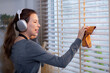 Excited young asian woman with headphones singing and dancing while dusting a window in living room, well-decorated room, joyful woman singing while cleaning in living room, lifestyle concept.