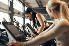 Fit Young Woman And A Group Of Female Friends In Sportswear Laughing While Riding Stationary Bikes During A Cardio Workout Session Together In A Health Club