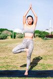 Fototapeta Las - young asian woman in sportswear doing tree position on her yoga mat at park, active and healthy lifestyle concept