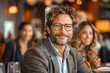 A cheerful, mature man with curly hair and glasses smiles warmly in a well-lit café setting among blurred individuals