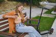 Side view happy fun young woman wear shirt casual clothes use mobile cell phone drink coffee sit on bench near e-scooter walk rest relax in spring city park outdoors on nature Urban lifestyle concept.