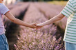 Friends hold hands closeup among lavender flowers in sunlight on summer day. Boy and girl walk in lavender field at sunset. Brother, sister have fun together. International Children's Day. Back view
