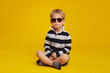 Cool little serious boy in striped shirt and trendy sunglasses feels confident while sitting crossed legged on floor against the yellow background