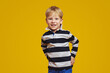 Cheerful little blonde child wearing striped shirt and keeping hands on waist while smiling for camera, against yellow background. Children studio portrait.