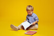 Positive curious schoolboy in casual clothes sitting on studio floor and reading notepad while smiling excitedly, isolated over yellow background.
