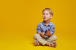 Thoughtful little boy in blue casual shirt sitting with crossed legs on studio floor and looking away while holding in hands a magnifier, isolated against yellow background. Free space for text