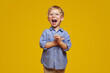 Little surprised child wearing striped blue shirt and stylish glasses clapping hands and yelling in amazement, standing isolated against yellow background.