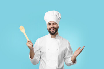 Sticker - Happy young chef in uniform holding wooden spoon on light blue background