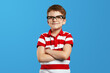 Smart little boy with wearing nerdy eyeglasses and striped red shirt smiling for camera while keeping hands crossed, standing isolated over blue background