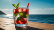 Cold refreshing mojito with slices of lime, mint, strawberries and pieces of ice with a red straw in the rays of the summer sun against the backdrop of the azure ocean