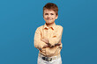 Adorable little kid boy wearing beige shirt smiling for camera while keeping hands crossed against blue background