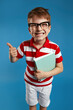 Close up portrait of smart wunderkind in nerdy eyeglasses and red striped shirt laughing and holding textbooks while showing thumb up gesture, standing isolated over blue background