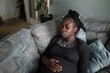 Black pregnant woman sitting on sofa in living room, sad African-American girl with big belly suffering from depression and crying, AI generated