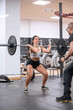© JoseIMartin - Fitness enthusiast performs a barbell squat in a gym under the watchful eye of her personal trainer, demonstrating proper form.
