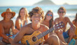 Handsome teenager boy playing Ukulele instrument on the seaside sandy ocean beach while spending funny time with friends. Active People, friends relations and summer vacation concept.