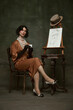© Lustre Art Group  - Elegant man in silky, old-fashion dress sitting by easel with self-portrait and drinking coffee against vintage studio background. Concept of self-expression, comparisons of eras, freedom, human right