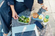 © okrasiuk - Top view business woman having lunch break, eating fast and healthy in front of laptop outdoors. Balanced diet lunchbox with fresh salad, sandwich, apple, water. Healthy eating habits and well-being.