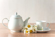 Herbal tea concept; white cup of tea, tea pot and bouquet of white chamomile flowers on a wooden table and white background; copy space