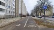 Asphalt sidewalks and a marked bike path are laid between the carriageway of the street with parked cars and residential buildings. Shrubs and trees stand on the snow-covered lawns. Cloudy weather