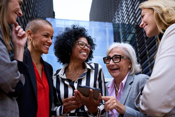 Professional business women laughing using and looking tablet digital display pc. Cheerful group of multicultural entrepreneur females conversing standing outside the office building having fun