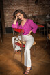 Beautiful young brunette woman in a purple blouse and white pants posing indoor against a brick wall. Portrait of a beautiful young brunette woman 