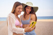 © CarlosBarquero - Two beautiful best friends looking at cell phone having fun on beach. Young happy women in swimwear point at mobile screen laughing. Couple of attractive generation z girls addicted to social networks