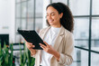 © Kateryna - Successful elegant hispanic or brazilian woman, successful employee of a company, female teacher, standing with documents folder in hands in a modern creative office, reading information, smiles
