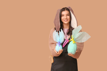 Sticker - Happy female hairdresser in bunny ears with chocolate Easter eggs and hairpins on brown background