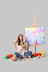 Sticker - Happy female artist in bunny ears with Easter baskets, toy carrots and easel sitting on grey background