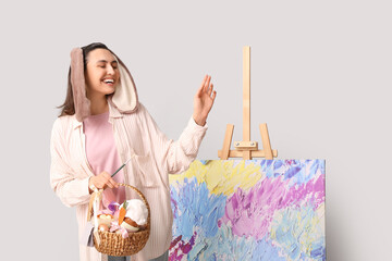 Poster - Happy female artist in bunny ears with Easter basket, brush and easel on grey background