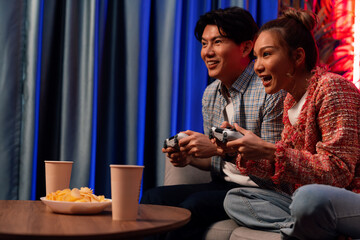 Sitting sofa of happy smiling couple playing video game control by joystick fronted snack and drinks competing challenge level with funny at modern neon blub light comfy living room at home. Infobahn.