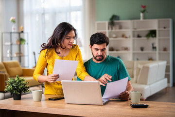 Young Indian couple checking mortgage or loan agreement, financial documents together, using laptop and calculator.