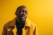 A black man in his 30s wearing a yellow coat crying exaggeratedly and overactingly on a yellow background