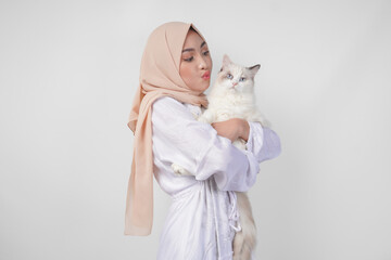 Wall Mural - Portrait of a beautiful young Muslim woman wearing white dress and hijab, smiling and holding a white ragdoll cat over isolated white background