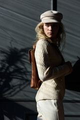 Poster - Studio portrait of beautiful woman with a curly blond hair holding brown backpack, posing on gray background.