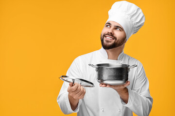 Canvas Print - Happy young chef in uniform holding cooking pot on orange background. Space for text