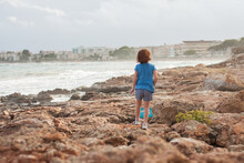 Girl While Walking Along The Pebble Beach To The Sea Against The Backdrop City. Rocky Coast At Sunny Afternoon. Mediterranean