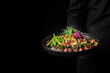 A person holds a plate of colorful, freshly prepared salad with grilled meat against a dark background. The vibrant dish contrasts beautifully with the backdrop