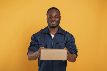Wall Mural - Delivery service worker with box. Handsome black man is in the studio against yellow background