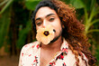 Dark-skinned man with curly long hair poses in nature, flower in mouth expressing confidence, non-conformity. Hawaiian shirt blends with green background, representing LGBT pride, ethnic diversity.