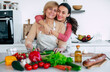 Smiling mature mother and her expressive lovely adult daughter cutting vegetables for a vegan salad together. Mid-adult woman and cute girl are preparing proper healthy meal on domestic kitchen