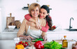 Smiling mature mother and her expressive lovely adult daughter cutting vegetables for a vegan salad together. Mid-adult woman and cute girl are preparing proper healthy meal on domestic kitchen