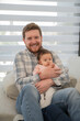 © Julio - Portrait of dad holding his baby daughter in his arms, It's daytime he looks at the camera sitting on the sofa.