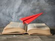 Closeup of a red paper plane on an open book with pages flying, embarking on the journey of knowledge and discovery.