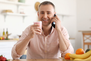 Wall Mural - Mature man with glass of fresh fruit smoothie talking by mobile phone in kitchen