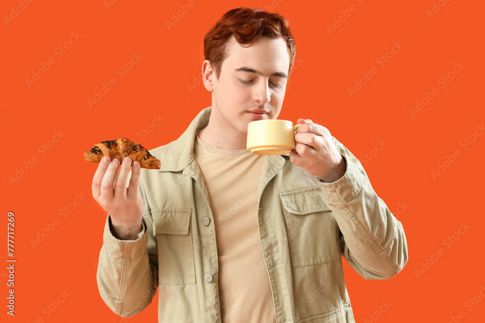Young man with cup of tea and croissant on orange background